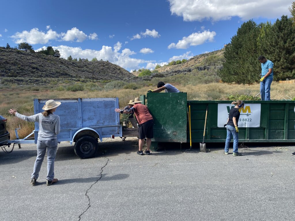 A community clean up event with a car with a trailer backed up to a dumpster. A group of people moving vegetation from the trailer into the dumpster.